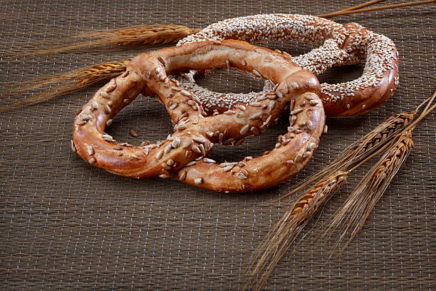 Two golden pretzels topped with sunflower and sesame seeds, arranged beside wheat stalks on a rustic woven kitchen mat in a warm, natural setting
