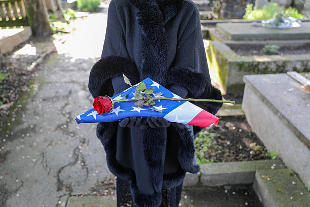 A mature woman, dressed in black mourning attire with a headscarf and black veil, standing solemnly in front of a tombstone at a cemetery. The scene conveys a sense of grief and respect, capturing the tradition of mourning and reflection at a final resting place. The soft lighting and somber expression evoke emotions of loss, remembrance, and reverence.