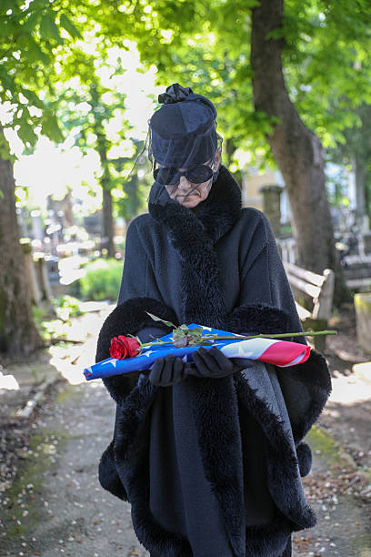 A mature woman, dressed in black mourning attire with a headscarf and black veil, standing solemnly in front of a tombstone at a cemetery. The scene conveys a sense of grief and respect, capturing the tradition of mourning and reflection at a final resting place. The soft lighting and somber expression evoke emotions of loss, remembrance, and reverence.