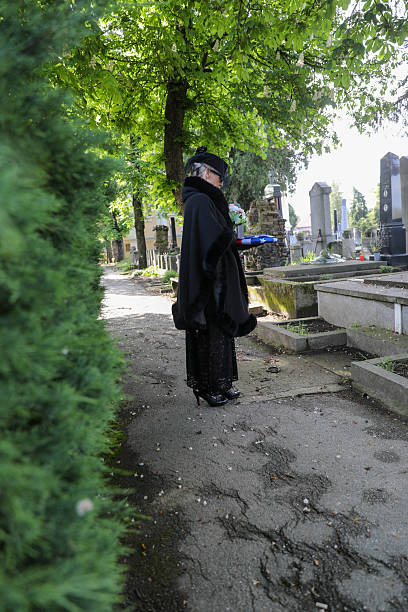 Elderly Mourning Woman In Black Traditional Attire Holding A Bouquet Of Flowers And A Folded Flag In A Quiet Cemetery, Symbolizing Grief, Respect, Remembrance, And National Honor During A Solemn Memorial Tribute.