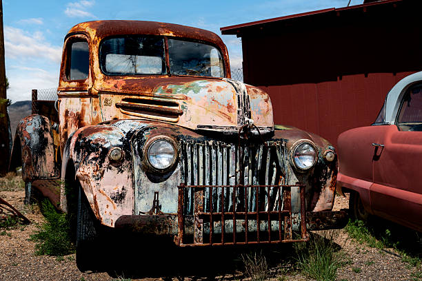 This abandoned antique pickup truck, painted in faded orange, sits in a desert junkyard along historic U.S. Route 66 in Arizona. A hand-painted '66' sign links the vehicle directly to the highway's cultural legacy and its role in mid-20th-century American travel and commerce. Weathered metal, peeling paint, and the surrounding landscape reflect the decline of roadside industry following the rise of the interstate system, leaving the truck as a quiet remnant of Route 66's past.