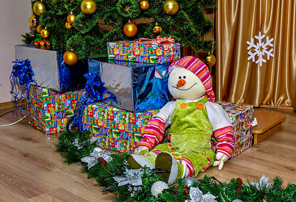 A snowman doll is sitting in front of a Christmas tree with wrapped presents