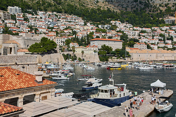 Dubrovnik. Croatia - November 20, 2025: High angle view of the Old Port in Dubrovnik, Croatia. Tourists walk on the pier with boats, city walls, and Mount Srd in the background.