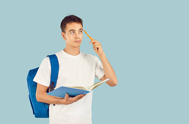 istock Young thinking brunette student boy holding a pen and textbook with backpack on blue background. 2247839032