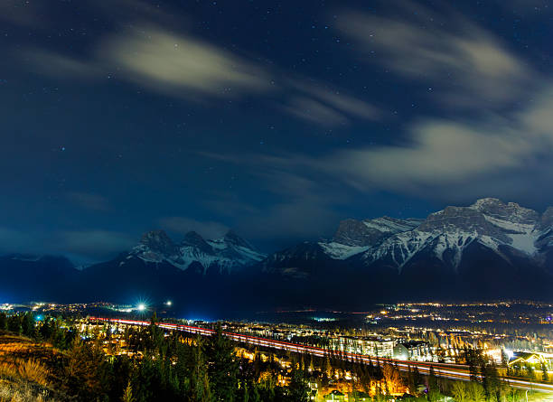 A view of the town of Canmore in Alberta, Canada, at night. The Three Sisters Mountains are visible on the left. The lights from the traffic on the Trans-Canada Highway are visible at the bottom.