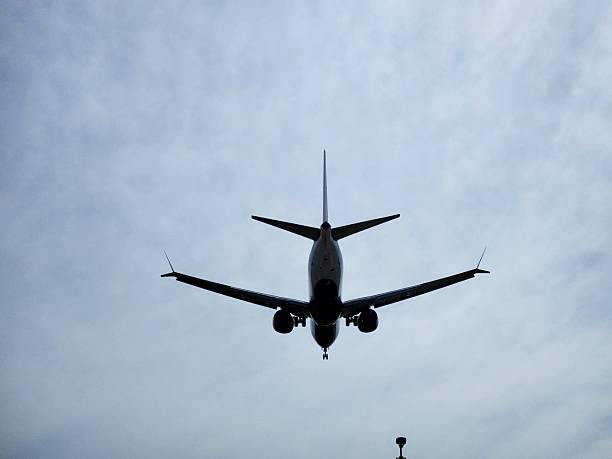 A commercial passenger jet captured from below while approaching the runway. The aircraft forms a dramatic silhouette against a cloudy sky, showcasing its landing gear and wings in detail. Perfect for themes related to aviation, travel, airports, transportation, and flight safety.