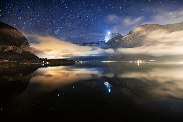 Clouds and stars above Lake Hallstatt with glowing mountain peak and reflections on calm water at night