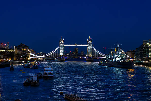 A wide shot of London's iconic Tower Bridge and the River Thames at night. The bridge is brightly illuminated with white lights outlining its Gothic towers and suspension cables, creating a striking contrast against the deep blue twilight sky. To the right of the bridge, the historic HMS Belfast warship is prominently docked, also lit up, its structure reflecting on the dark, rippling water of the Thames. Numerous smaller boats are visible on the river in the foreground, with their own subtle lights and reflections. The city skyline on both banks features various buildings with illuminated windows, adding to the urban glow. The overall scene conveys the majestic and vibrant atmosphere of London after dark, showcasing its architectural heritage and bustling river life.