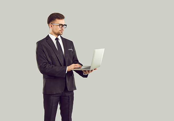 istock Serious businessman standing isolated on gray background and working on laptop computer 2247849806