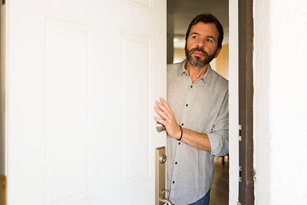 Mature man peeking cautiously and looking outside while opening the front door of his home, feeling alert or curious