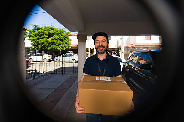 istock Delivery man holding cardboard box at front door, offering package with a smile from a peephole perspective 2247859859