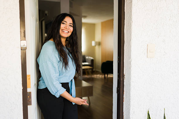 istock Young woman smiling and gesturing with hand, standing at open front door, inviting visitors inside her house 2247860442