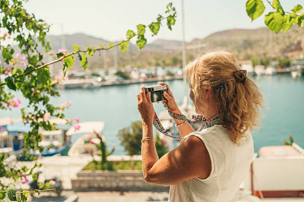 Side view of a woman taking photos of boats and coastal landscape from a balcony decorated with plants overlooking a marina.
