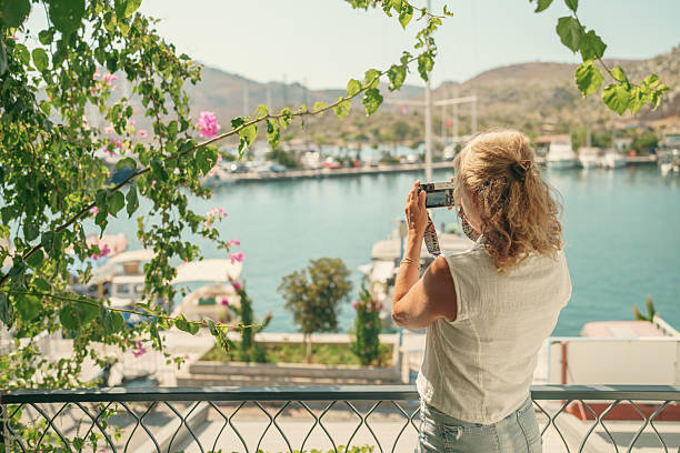 Side view of a woman taking photos of boats and coastal landscape from a balcony decorated with plants overlooking a marina.