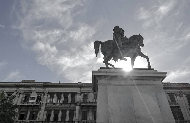 A powerful low-angle, black and white photograph of an equestrian statue, likely depicting a historical figure, silhouetted against a dramatic sky filled with wispy clouds.