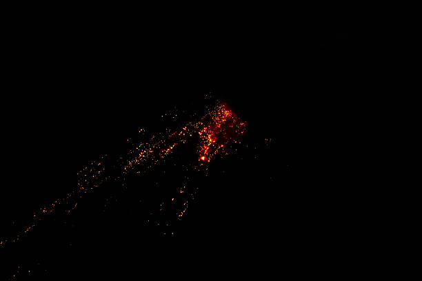 An aerial, dark-night view captures the dramatic, incandescent glow of an eruption on Barren Island volcano, located in the Andaman Sea. Bright red-orange hot magma and scattered lava flows illuminate the pitch-black surroundings, appearing as a fiery, abstract cluster against the darkness. The image focuses on the central point of volcanic activity, highlighting the raw power and beauty of a geological event.
