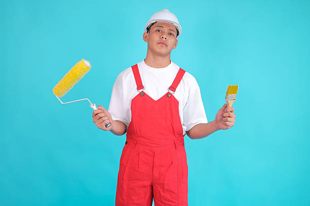istock Confused Handyman Holding Paint Roller and Brush, Questioning Renovation Project on Blue Backdrop 2247881416