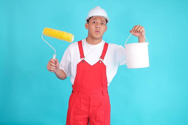 istock Painter in Hard Hat and Overalls Showing Brush Roller and Paint Bucket Against Blue Background 2247881423