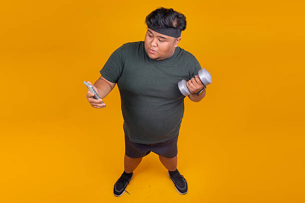istock Plus-sized Man with Dumbbell Checking Phone for Workout Motivation Isolated on Yellow Backdrop 2247884855