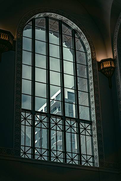 This image captures a moody and atmospheric urban scene viewed through an ornate arched window with intricate metalwork and stone detailing. The reflection of a cityscape, characterized by modern buildings, contrasts with the historic architectural style of the window, creating a blend of old and new. The subdued lighting and monochromatic tones evoke a contemplative and timeless mood, ideal for editorial, architectural, or travel-related commercial use.