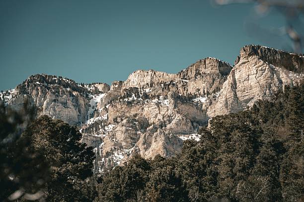 An expansive alpine panorama reveals the rugged upper ridges of the Teton Range, still blanketed with late-spring snow. Sheer rock walls rise into a sea of shifting cumulus clouds and deep blue sky, capturing the raw geometry of high mountain wilderness. Below, sparse conifer clusters trace the lines of ancient avalanches across the snowfields. The wooden snow fence in the lower corner provides a human scale to the otherwise untouched landscape. This image embodies the wild serenity of high-altitude Wyoming — a study in contrasts between earth, sky, and silence.