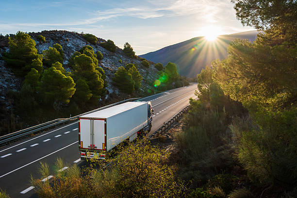 istock Refrigerated truck driving along a mountain road surrounded by forests, with sunlight breaking over the horizon above the peaks. 2247890785