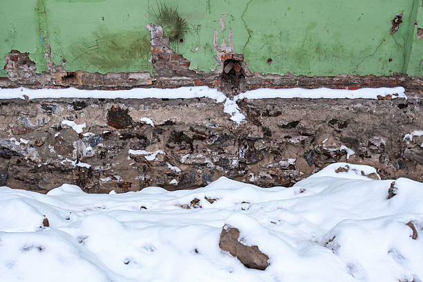 Old damaged wall with green peeling paint and exposed red bricks stands above a rough stone foundation covered with white fresh snow in winter creating a grunge abstract texture background