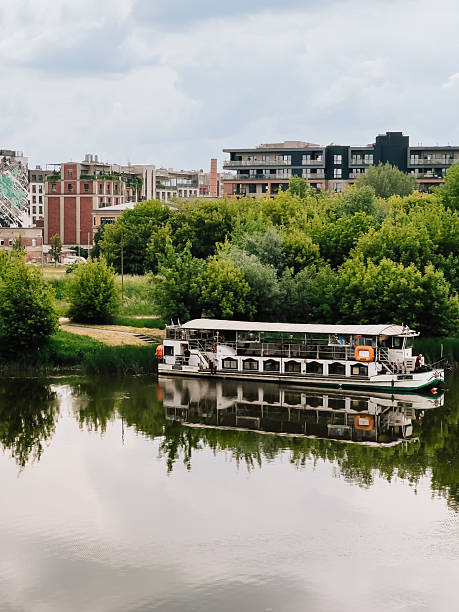 Passenger ferry moored on Vistula River reflecting on calm water surface with green trees and modern residential buildings on opposite bank on cloudy summer day in Krakow, Poland. Zablocie, Krakow