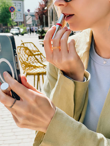 istock Professional woman checking smartphone while touching up red lipstick on urban sidewalk, reflecting modern workplace multitasking and personal grooming. Girl applying red lipstick during her break 2247900592