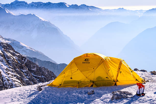 A vibrant yellow tent stands on a snowy mountain peak, surrounded by rugged, snow-covered terrain. Majestic, misty mountains in the background create a serene winter landscape, evoking adventure and solitude in the high alpine environment.
