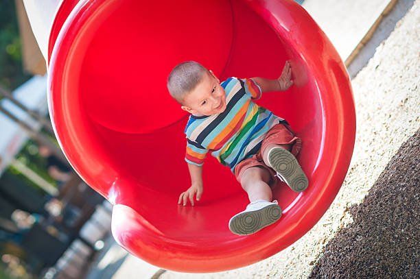 A young boy in a colorful striped shirt is sliding down a vibrant red slide in a playground. The scene captures his excitement as he enjoys this sunny outdoor activity.