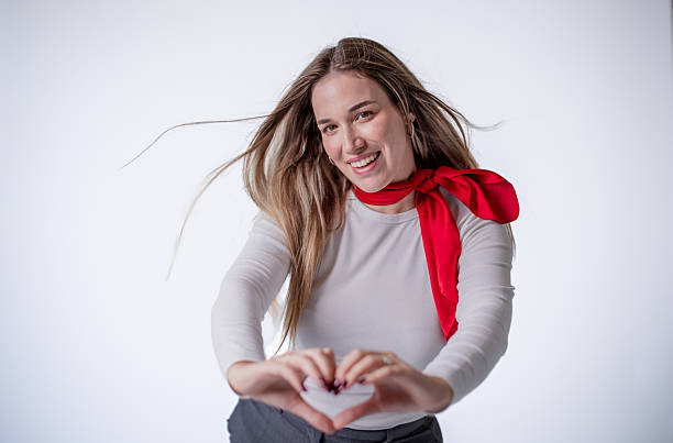 A cheerful woman wearing a gray top and a red scarf creates a heart shape with her hands. She is smiling joyfully, standing in front of a plain white background, radiating happiness.