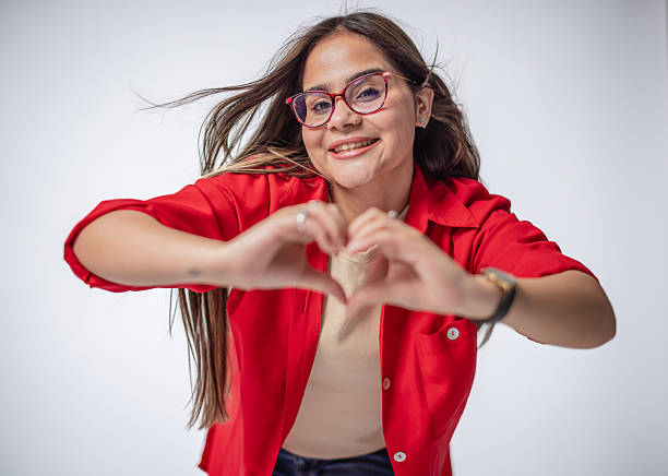 A cheerful woman with long hair smiles brightly while forming a heart shape with her hands. She wears a red shirt and stands against a plain white background, radiating positivity.