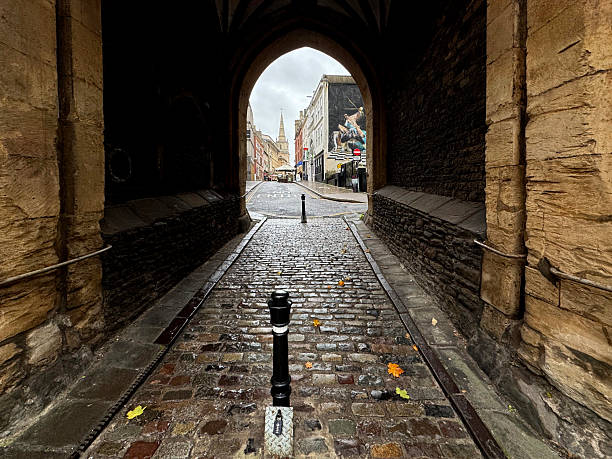 A view through a dark, stone archway looking out onto Christmas Street, in Bristol UK, a cobblestone street. The archway frames the bright street scene beyond it, creating a strong contrast. The street is wet and lined with various buildings. A black bollard is prominent in the immediate foreground, with another further down the street.