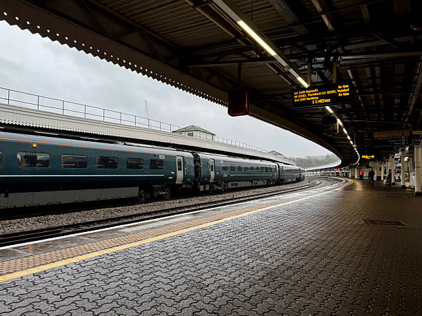 A dark green passenger train is stopped at Bristol Temple Meads station platform on an overcast day. The platform surface is made of textured, grey paving stones. Overhead canopies with integrated lighting cover the platform area.