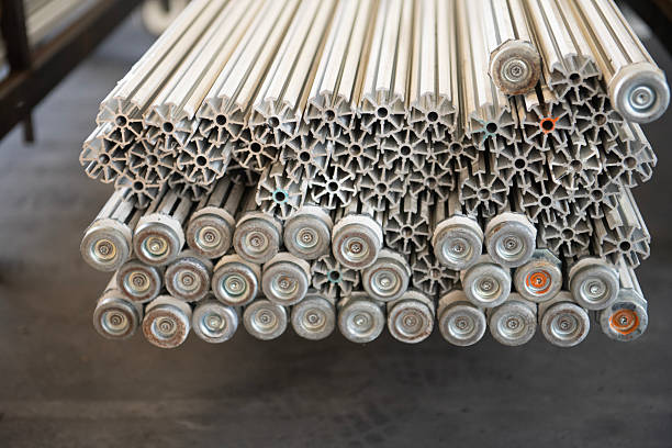 Stack of cylindrical aluminum poles with end caps, arranged closely together in a workshop or storage area; industrial metal texture, repeating geometric pattern, shallow depth of field.
