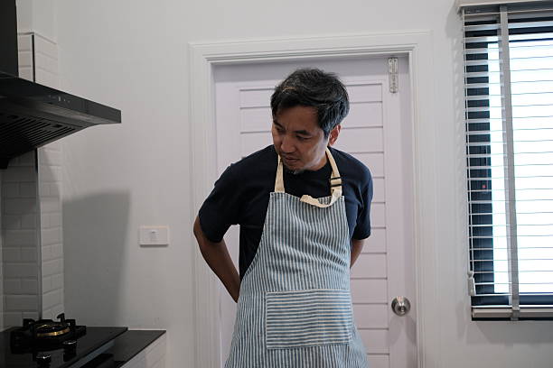A man in a blue shirt and a striped apron stands in a kitchen. He is looking at something behind him