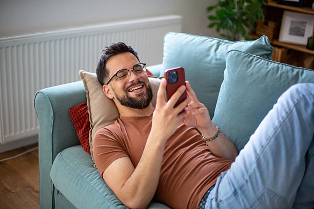 istock Young man laughing while relaxing on couch with smartphone 2247953211