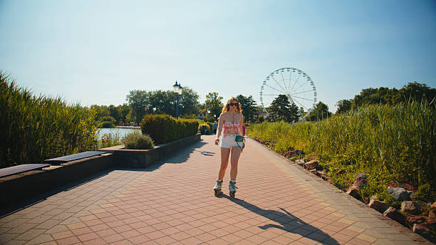 Caucasian woman joyfully roller skating along a sunlit path with a retro cassette player. Surrounded by greenery and a Ferris wheel in the background, she enjoys a leisurely outdoor experience on a clear day.