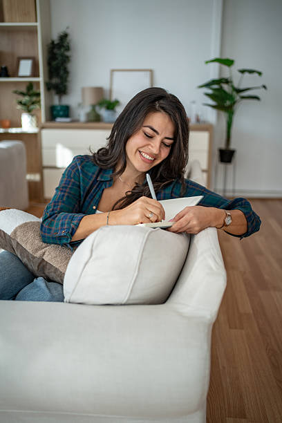 istock Young woman writing journal on couch at home 2247963121