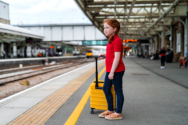 A young girl stands on a train platform, holding a yellow suitcase. She gazes at the tracks, waiting for her train on a cloudy day.