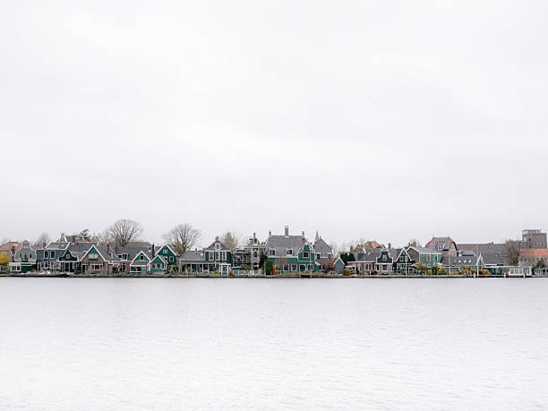 Panoramic view of traditional green wooden houses along the Zaan river in Zaandijk, Netherlands. Historic Dutch waterfront skyline featuring Gortershoek architecture under an overcast sky.