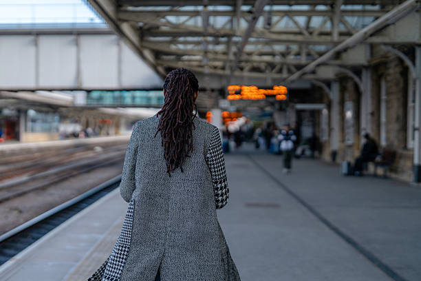 A woman with long hair walks along a train platform. Signs show train information while other travelers await their journeys in the background.