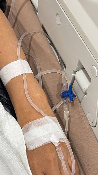 Close-up of a person’s arm with an IV cannula and transparent tubing secured with medical tape, receiving a saline infusion while resting on a hospital bed or clinic chair.