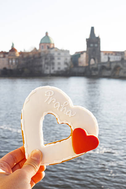 Hand holding a heart shaped gingerbread cookie with praha text, showing love for the city with charles bridge in blurred background