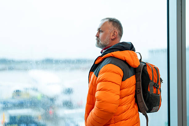 A man stands by the window in an airport terminal, wearing a bright orange jacket. He looks out over the tarmac, waiting for his flight during a cloudy day.