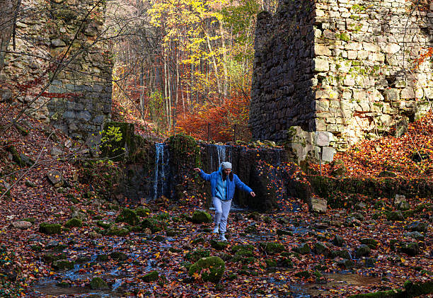 A person balances on moss-covered stones in a shallow stream beneath cascading water, framed by bright autumn trees and weathered stone walls. The scene captures movement, seasonal atmosphere, and natural exploration.