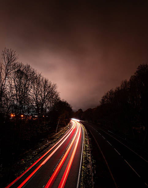 A stunning series of long-exposure photographs taken from an overpass, capturing the light trails of vehicles on a curving, dark dual carriageway (motorway/main road) at night. The slow shutter speed creates dynamic streaks of red (taillights) and bright white (headlights), emphasizing speed, movement, and the flow of traffic. The dark, tree-lined edges and the contrast with the low, cloudy sky enhance the moody, isolated, and energetic atmosphere of nighttime transport.