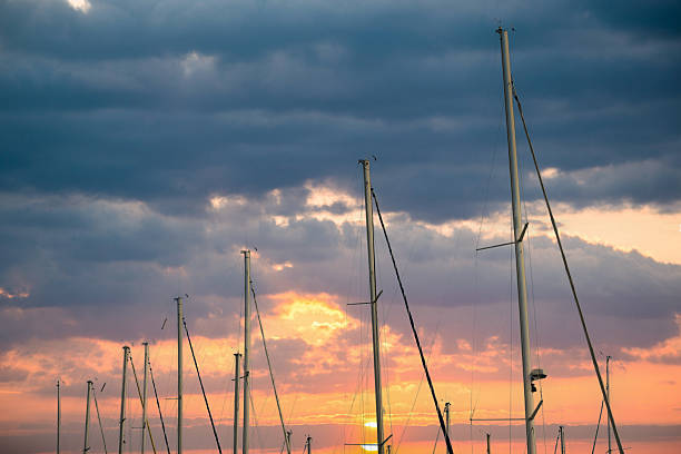 Sailboat masts silhouetted against a dramatic twilight sky of orange and blue, reflections on calm marina waters creating a tranquil, moody nautical evening scene