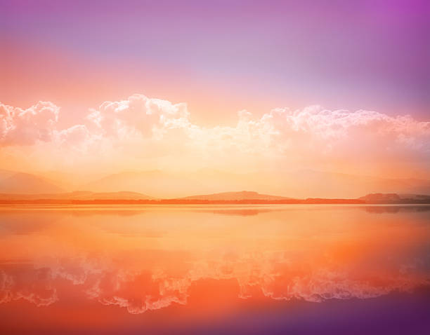 Serene lake mirroring a vibrant sunset sky with soft clouds and distant rolling mountains, warm gradient of orange, pink and purple creating a tranquil, reflective landscape